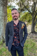 Photo of author Alexander Schneider. The author is standing in a clearing among trees, wearing a casual three-piece suit with a boutonnière.