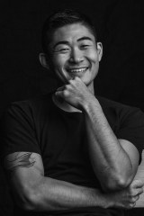 Black and white photo of artist Yoshino from the waist up. The artist is wearing a black shirt, and has one arm crossed in front with the other elbow sitting on that arm and raised up to his chin. He is smiling into the camera.